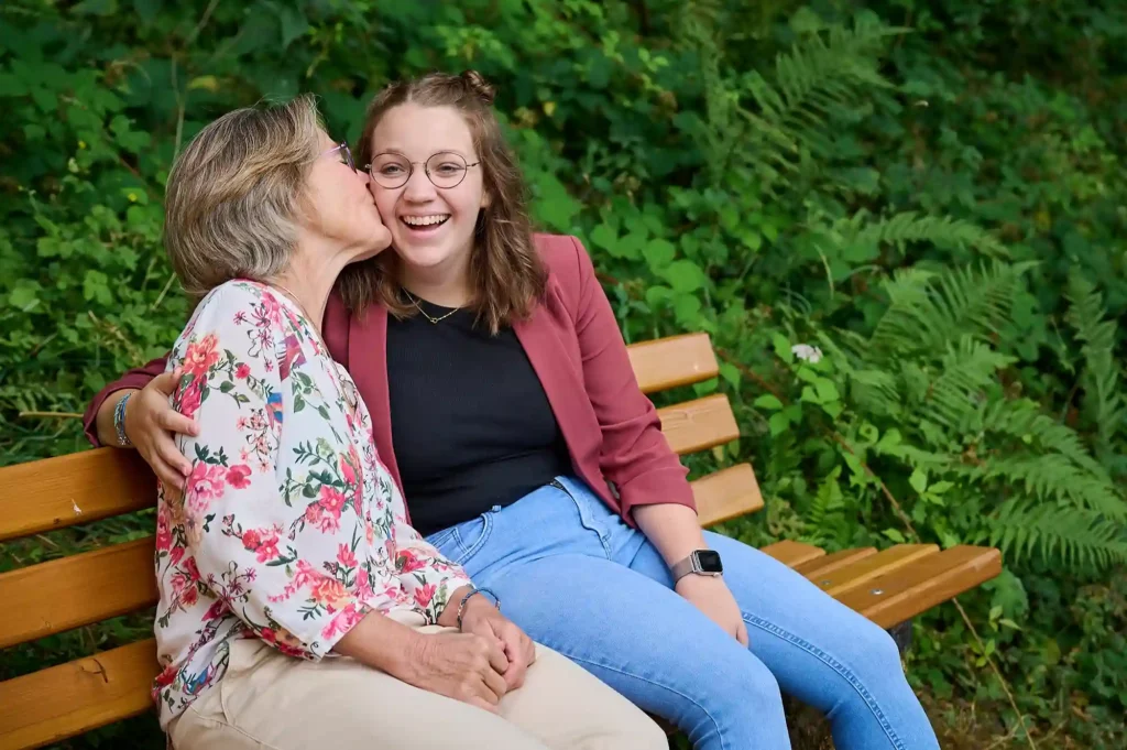 Eine Mutter küsst ihre Tochter, beide sitzen lächelnd auf einer Bank